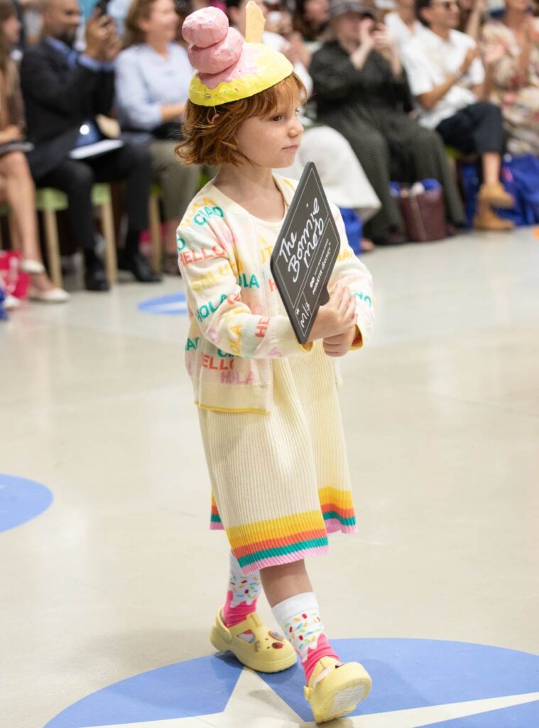 A child holding a sign on a stage wearing a dress and ice cream hat 