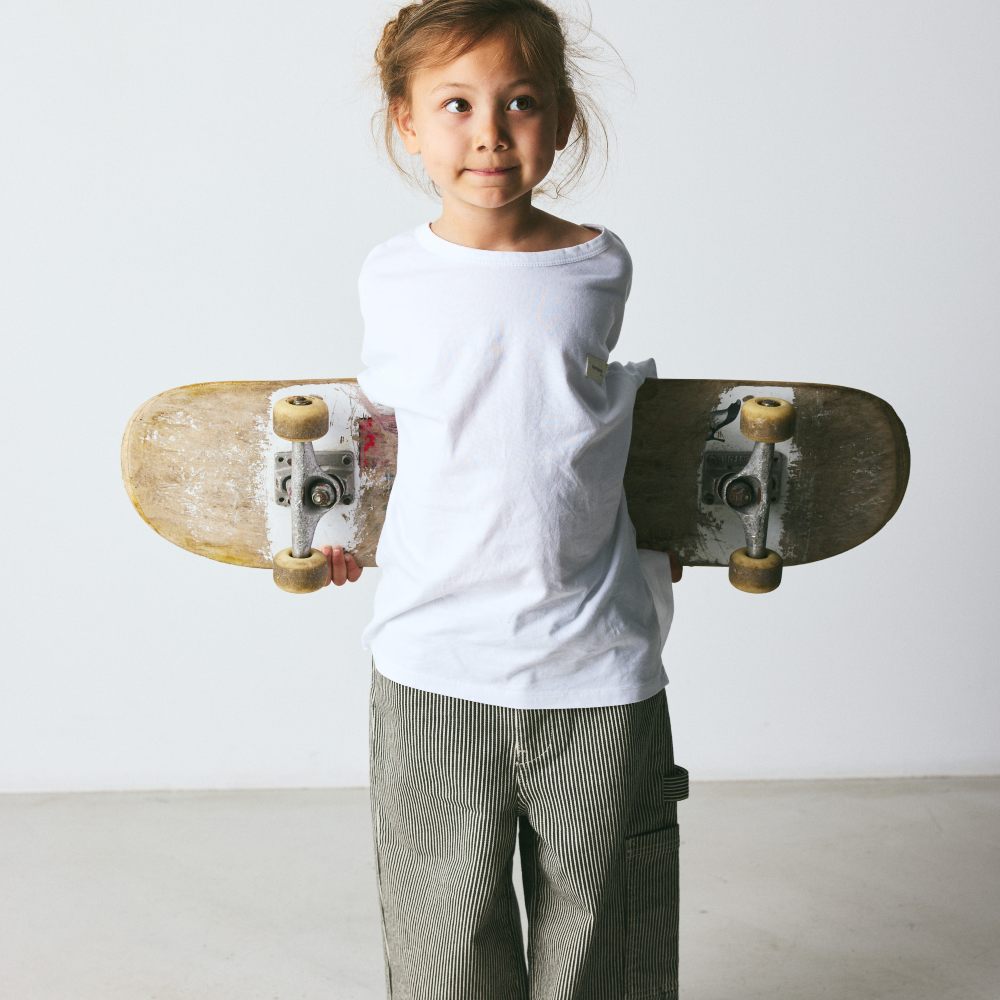 A girl wearing a top and trousers holding a skateboard behind her back 