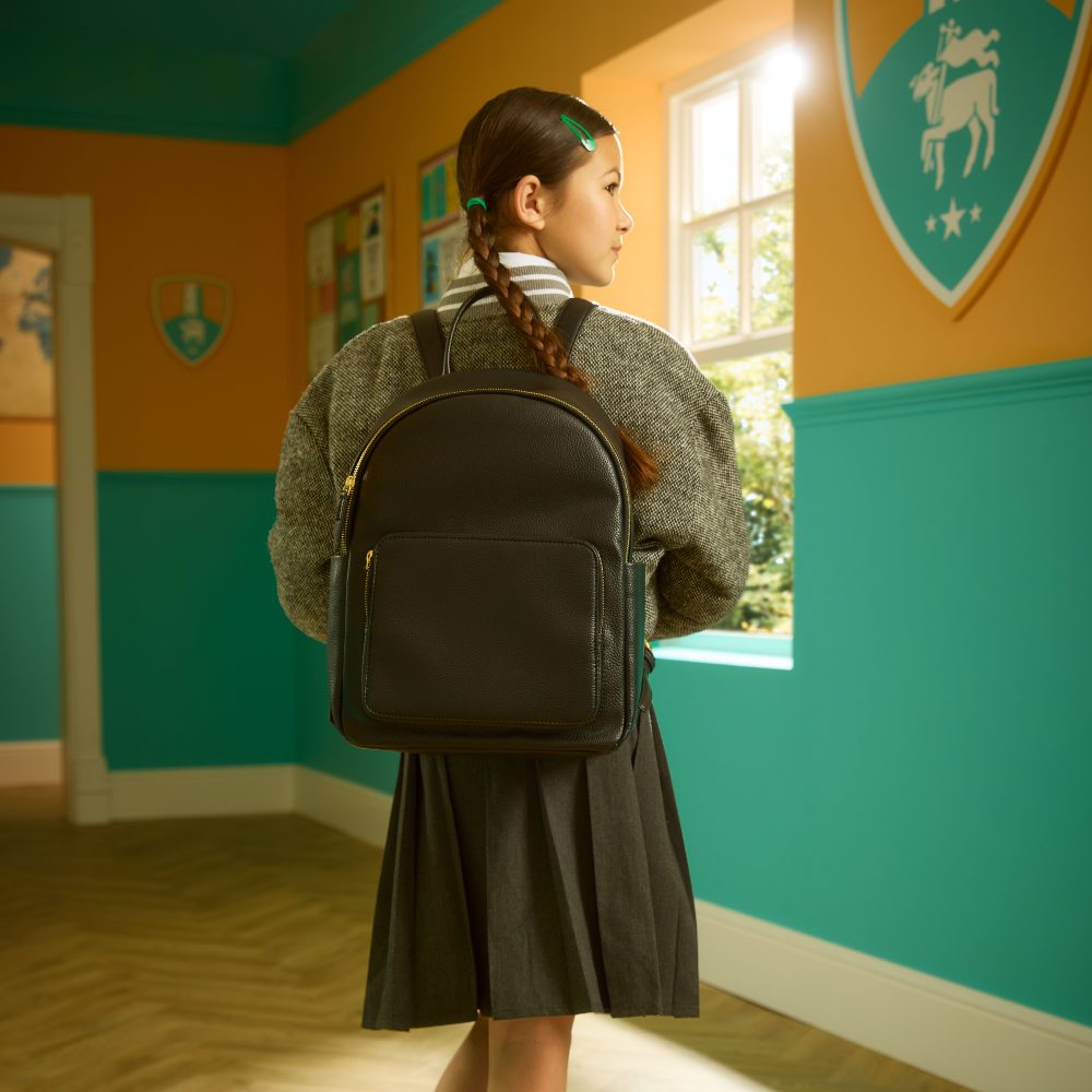 A girl stood in a corridor in school uniform and a black school bag by Clarks 