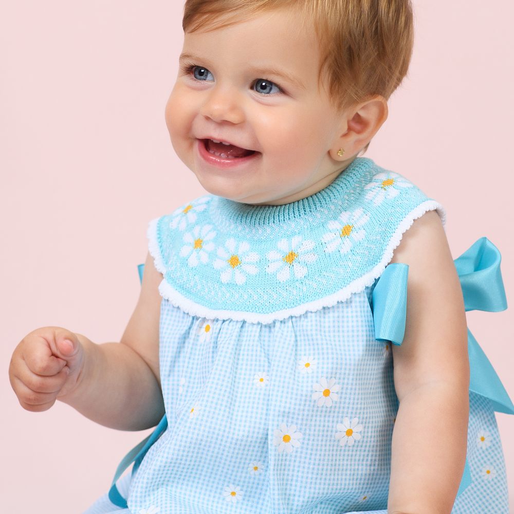 A baby smiling wearing a blue summer dress with daisy embroidery 