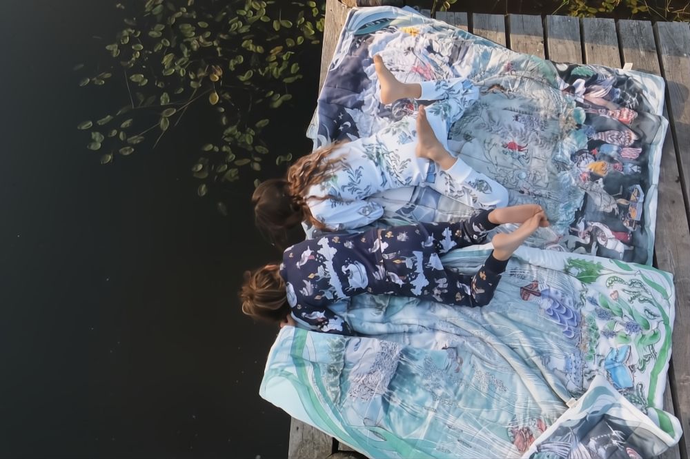Two children in pyjamas lying on printed blankets on a jetty and leaning down to look in the water 
