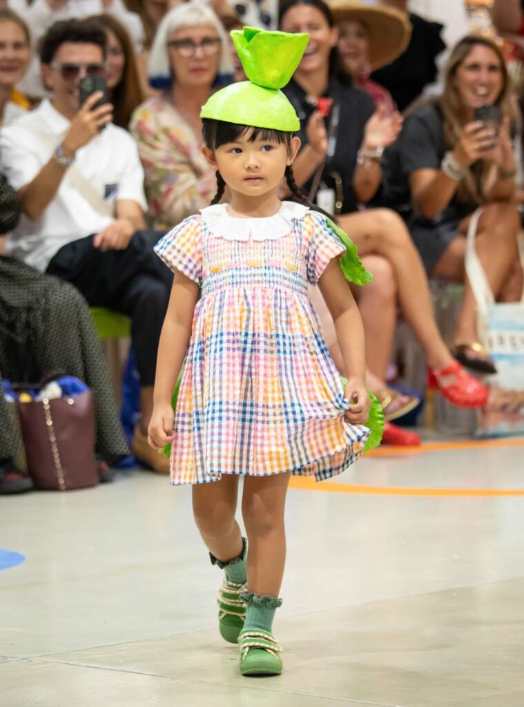 A child in a summer dress, green shoes and a hat walking in a fashion parade 