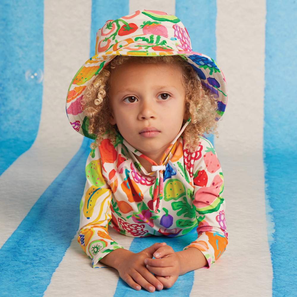 A child lying on their front on a blue and white striped background wearing a colourful fruit print sun hat and top 