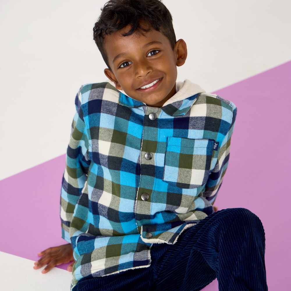 A smiling boy sat on the floor wearing a blue checked shirt and blue trousers 
