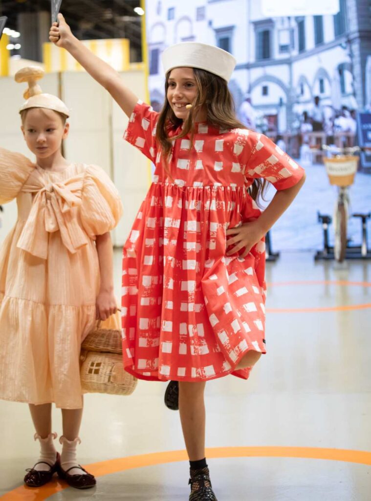A girl stood on a stage with her arm and leg in the air wearing a red and white check dress 