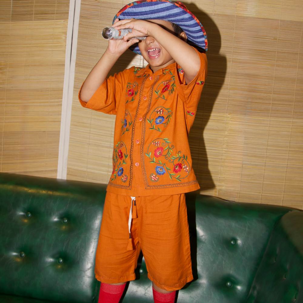 A boy stood on a green leather sofa wearing a hat and orange shorts and shirt holding a telescope to his eye