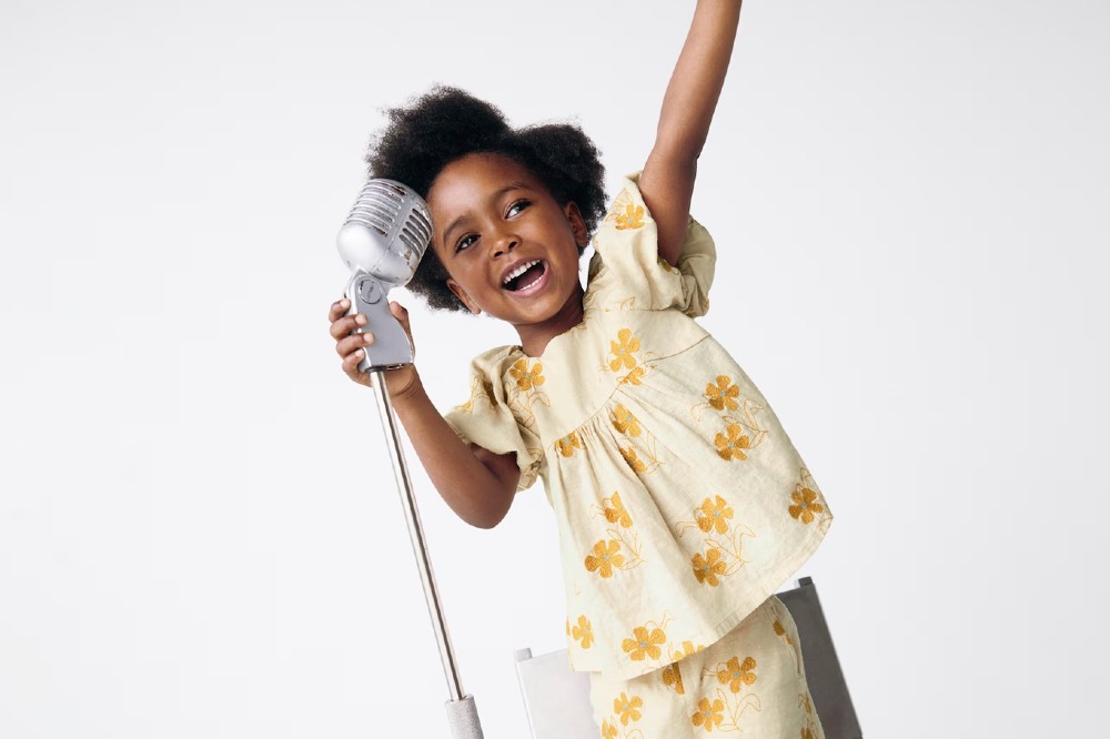 A girl holding her arm in the air holding a microphone and wearing a white top and trousers with yellow flower embroidery by M&S