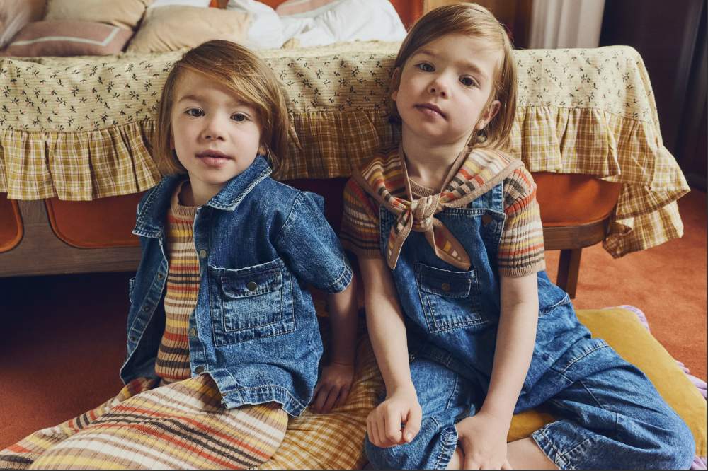 Two children sat on the floor at the foot of a bed wearing knitted and denim outfits 