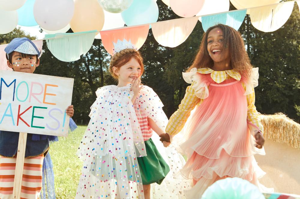 Three children stood outside in party and fancy dress clothes surrounded by balloons