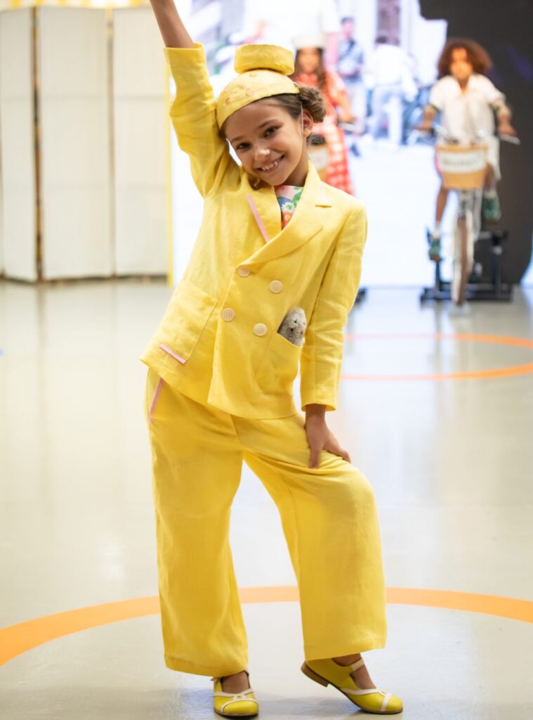 A girl in a yellow trouser suit and hat stood on a stage 
