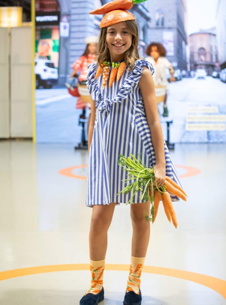 A girl in a blue and white striped dress and carrot hat holding a bunch of carrots 