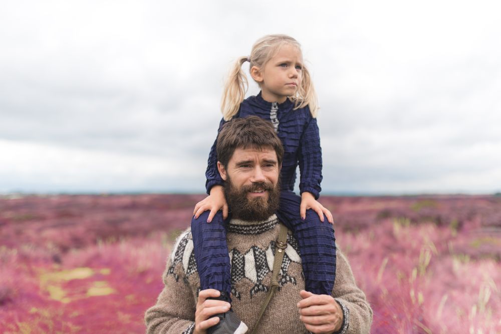 A child in a blue Petit Pli outfit sat on a man's shoulders outside on heather covered ground