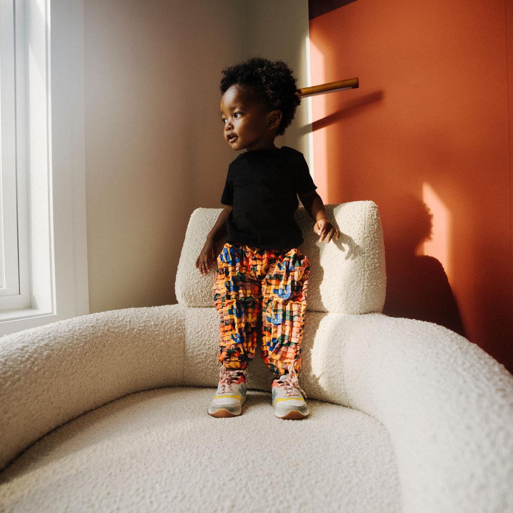 A child stood on a large white chair in a room with orange walls wearing colourful trousers and a black top 
