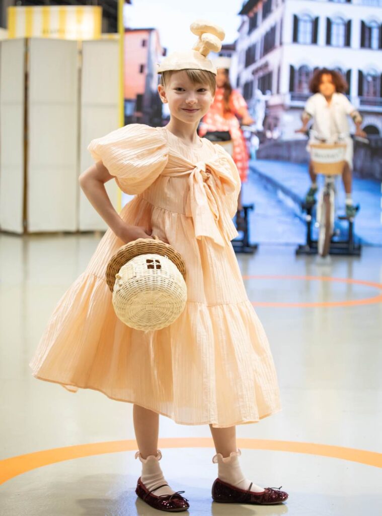A girl in a peach coloured full dress and hat holding a bag 