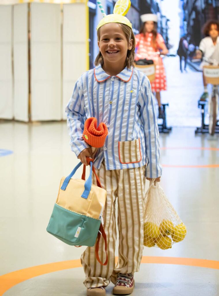 A boy in a striped shirt and trousers holding bags in one hand and a net of lemons in the other 