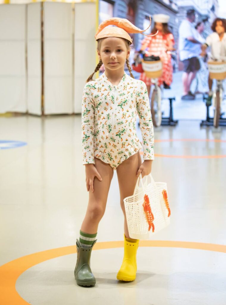 A child in a swimming costume and odd coloured wellies holding a bag 