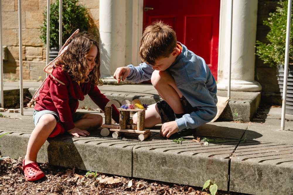 Two children sat on the front steps of a house playing with the Natural Blocks Kit by The Den Kit Company