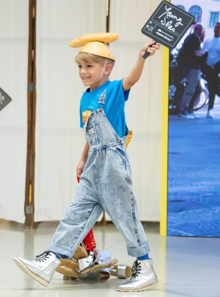 A boy in dungarees and a blue T-shirt holding up a sign and carrying a bunch of shoes 