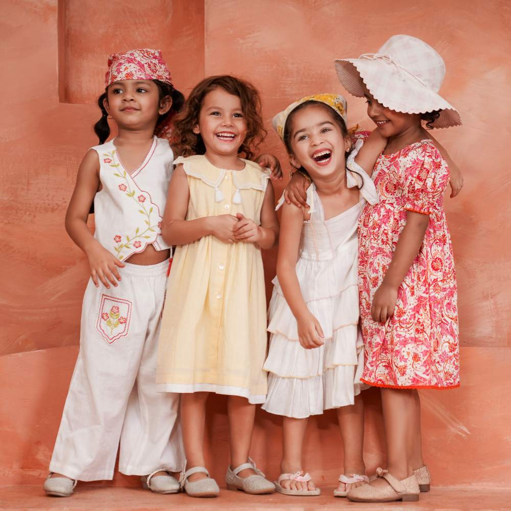 Four smiling girls in summer dresses stood against a pink background 
