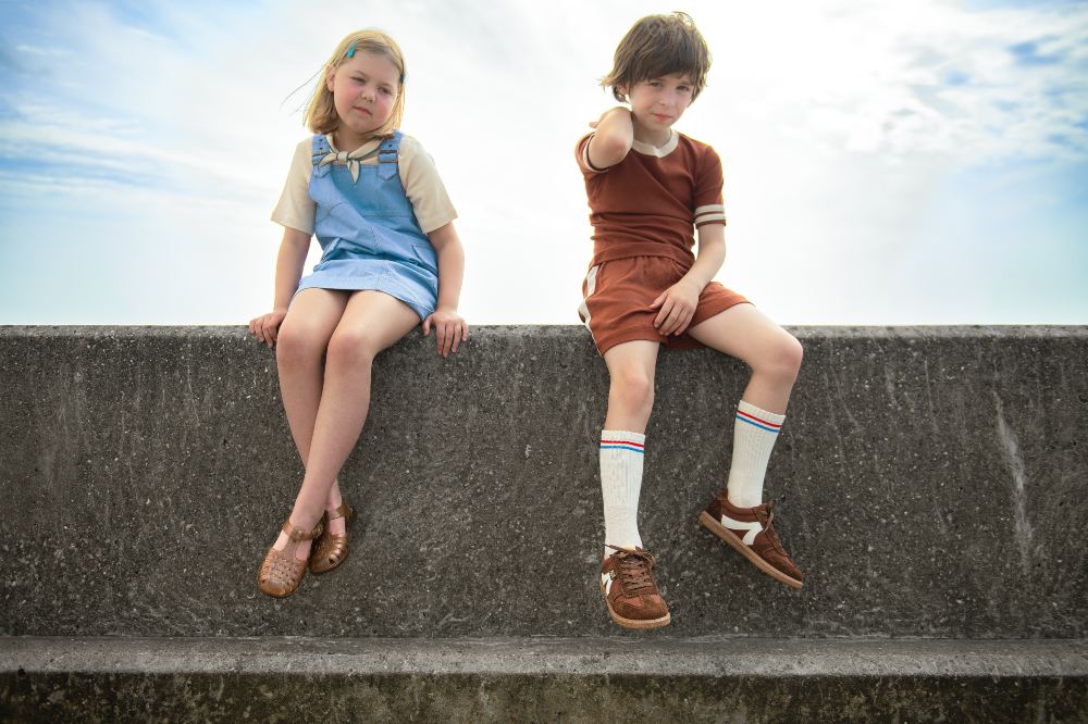 Two children sat on a sea wall wearing summer outfits and shoes by Young Soles