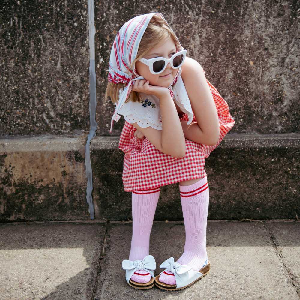 A girl sat on a wall wearing a headscarf, white sunglasses, a red check dress and socks and sliders 