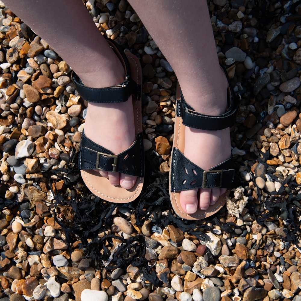 A child's legs and feet show stood on a pebble beach with seaweed wearing black summer sandals 