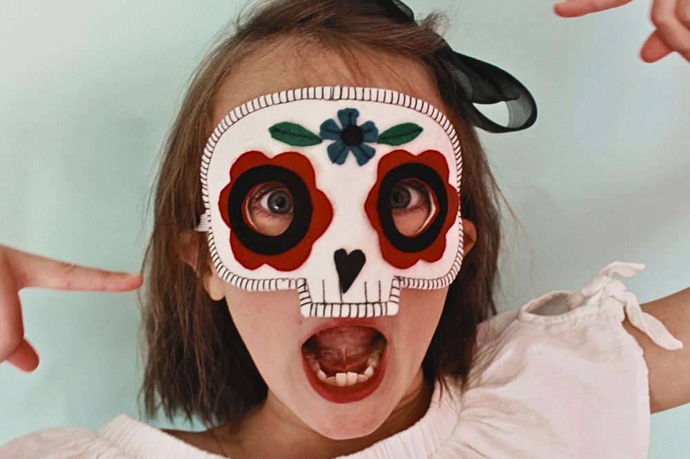 A young girl wearing a colourful skull mask from the A is for Alice Halloween Collection