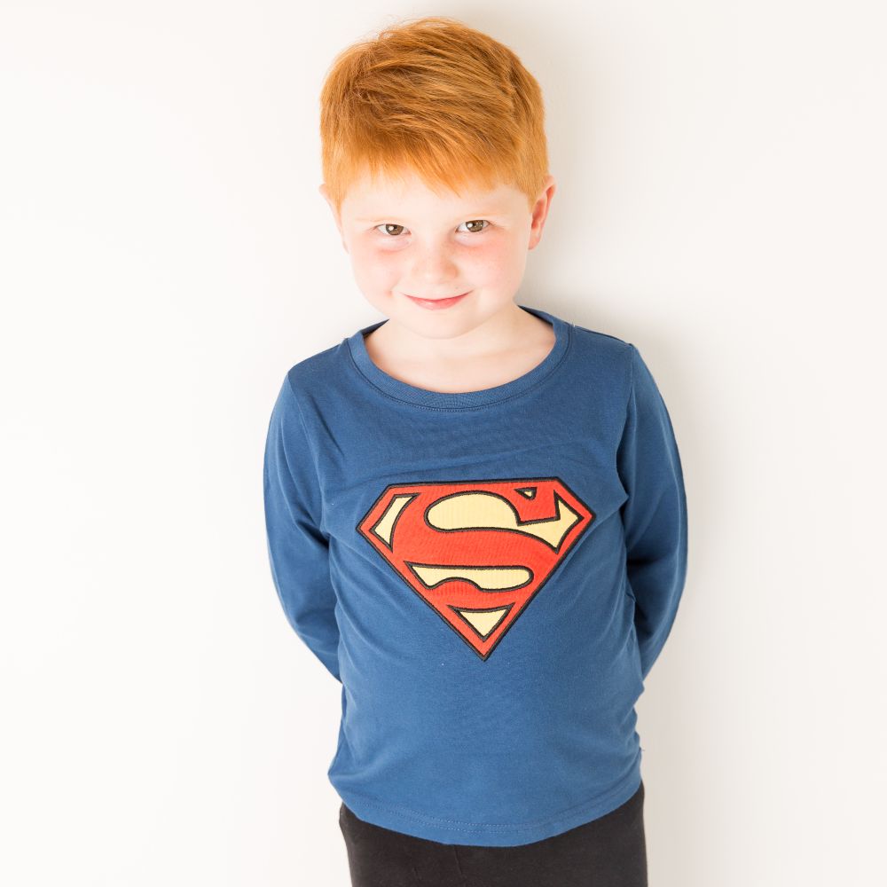A boy with ginger hair stood against a white wall wearing a blue top with the Superman logo by Blade & Rose 