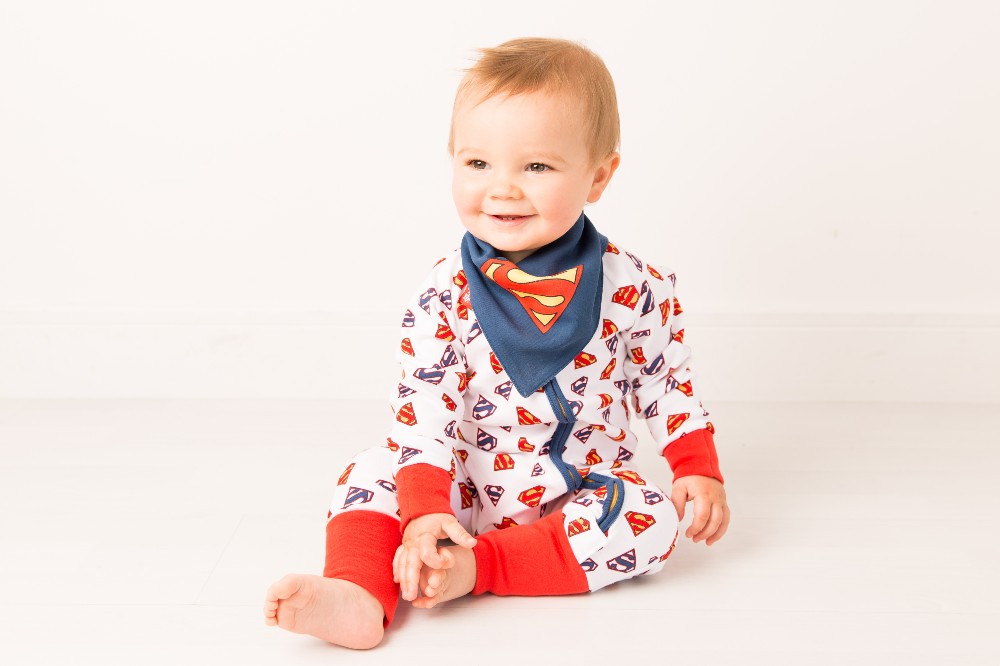 A baby sat on the floor wearing a Superman romper and bib by Blade & Rose