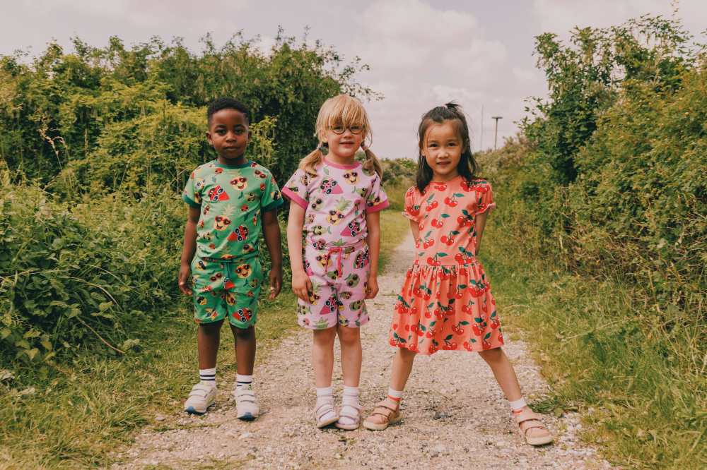 Three children dressed in brightly coloured summer outfits stood outside on a pathway