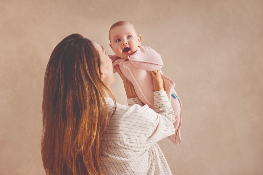 A woman holding up a young baby wearing an oatmeal coloured sleep swaddle by Love to Dream