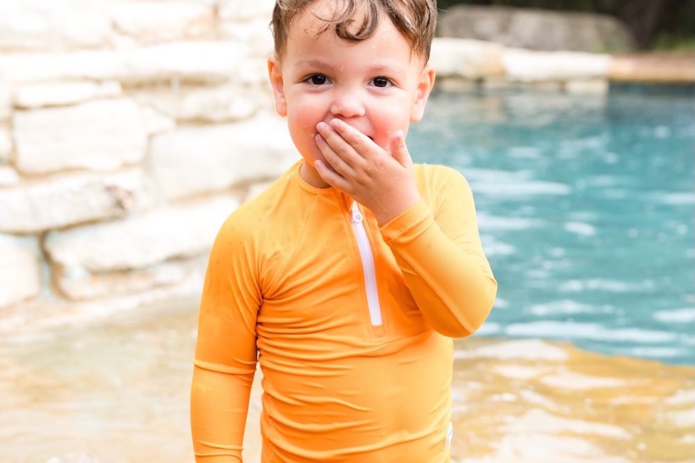 A boy stood outside in a pool wearing a bright orange UV rash vest by Splash About