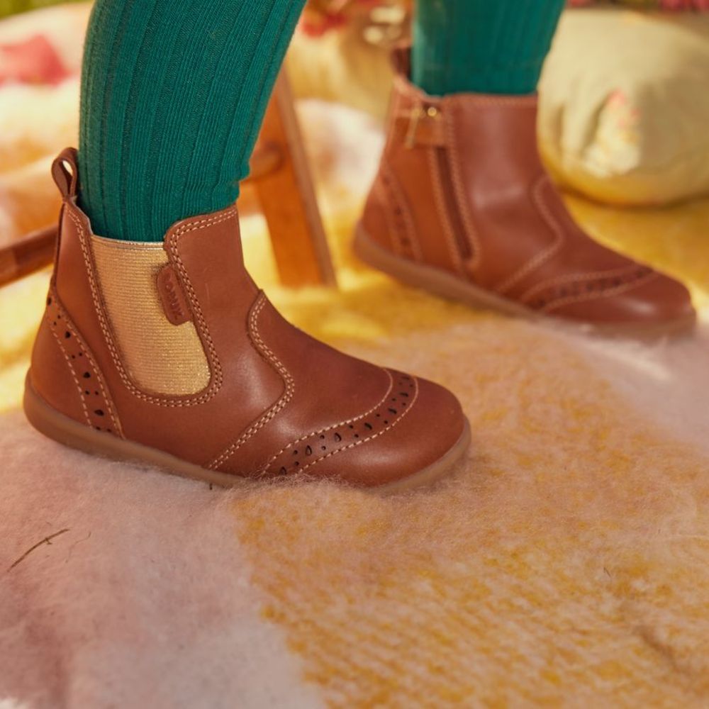 A child's feet shown stood on a pink blanket wearing brown boots with gold detailing by Bobux 