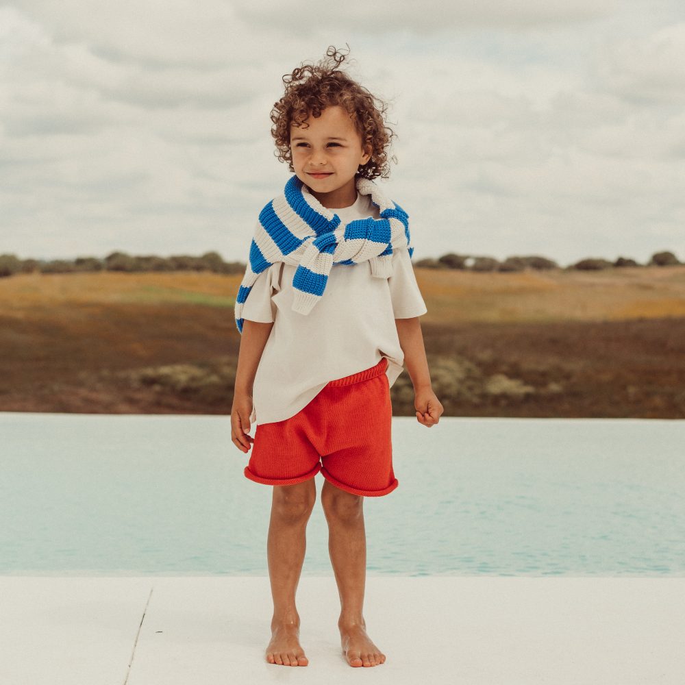 A young boy stood outside in red shorts and a T-shirt with a blue and white striped jumper over his shoulders 