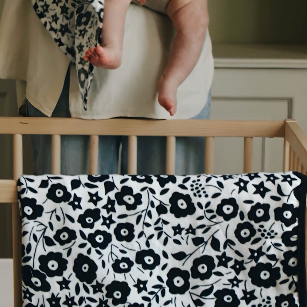 A person holding a baby beside a cot with an Etta Loves x Liberty blanket draped over it 