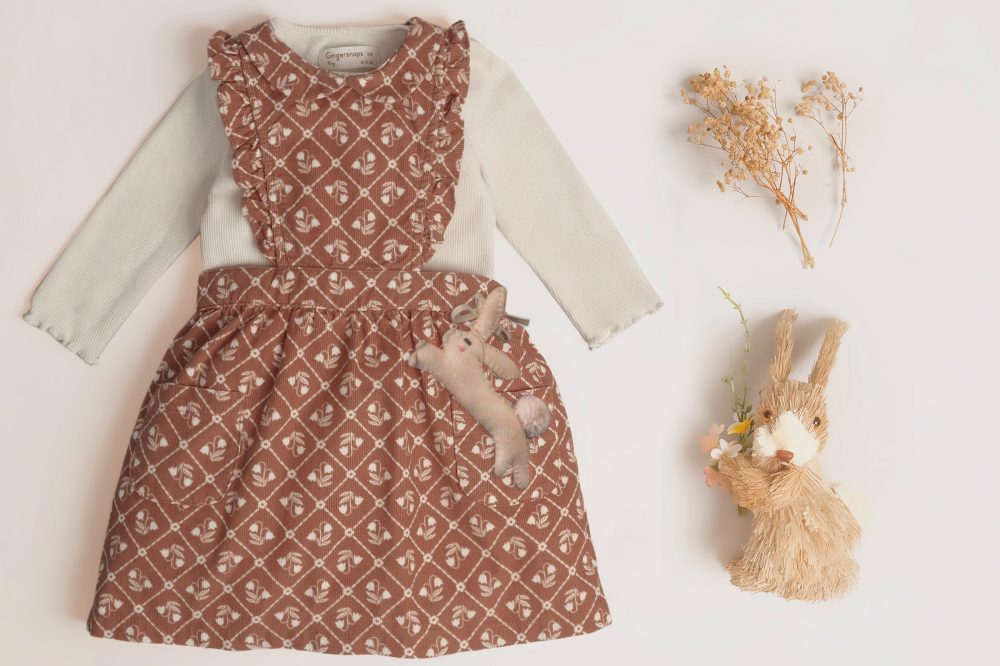 A baby girls dress and top by Gingersnaps displayed on a white background beside a toy rabbit and dried flowers