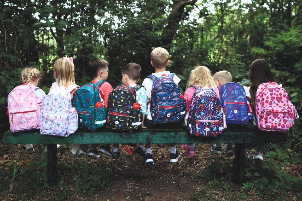 A row of children sat on a bench outside with their back to the camera showing backpacks by Roamie