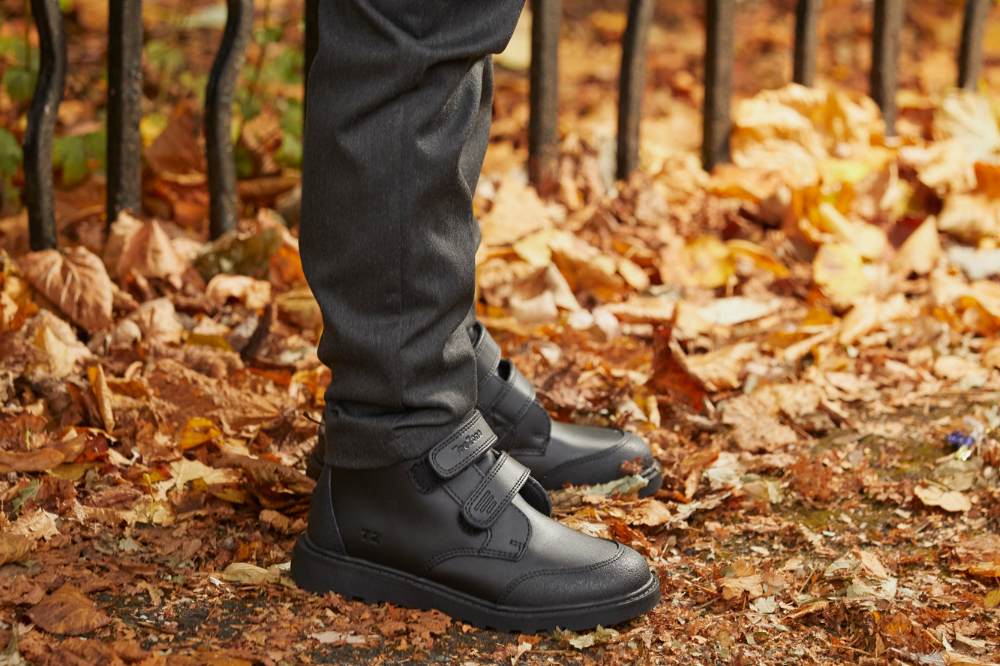 A child's legs shown stood outside in leaves by a fence wearing school trousers and black school shoes by ToeZone
