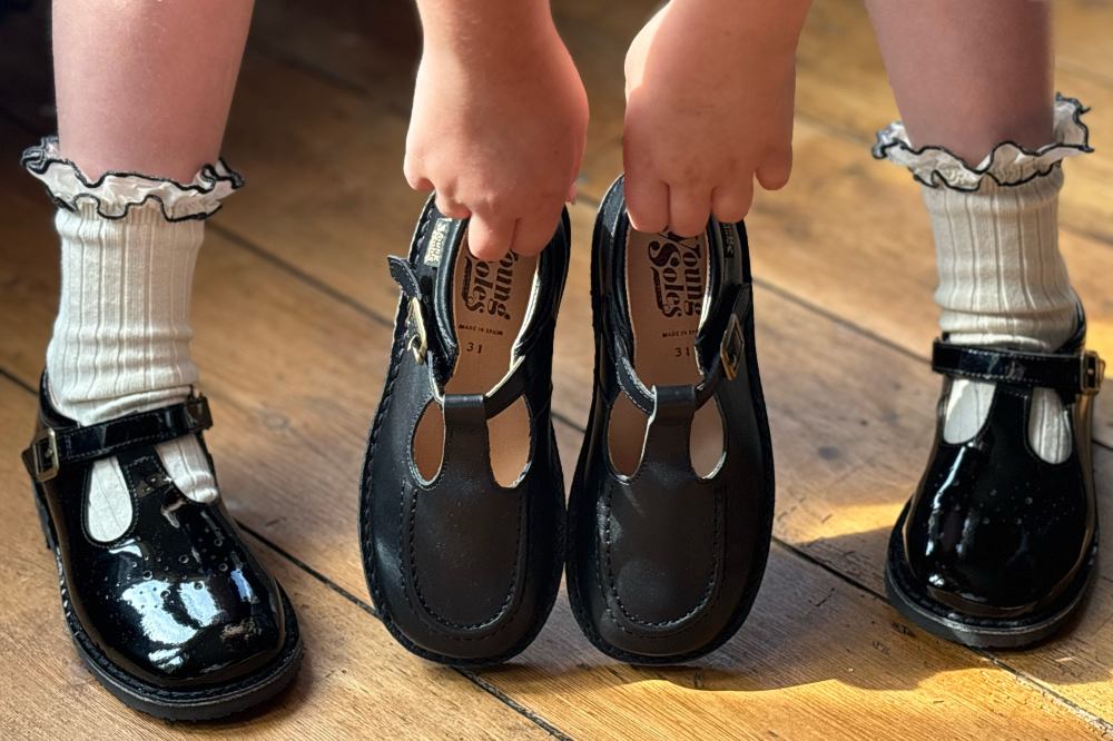 A child's hands shown holding a black pair of T-Bar shoes by Young Soles between her feet