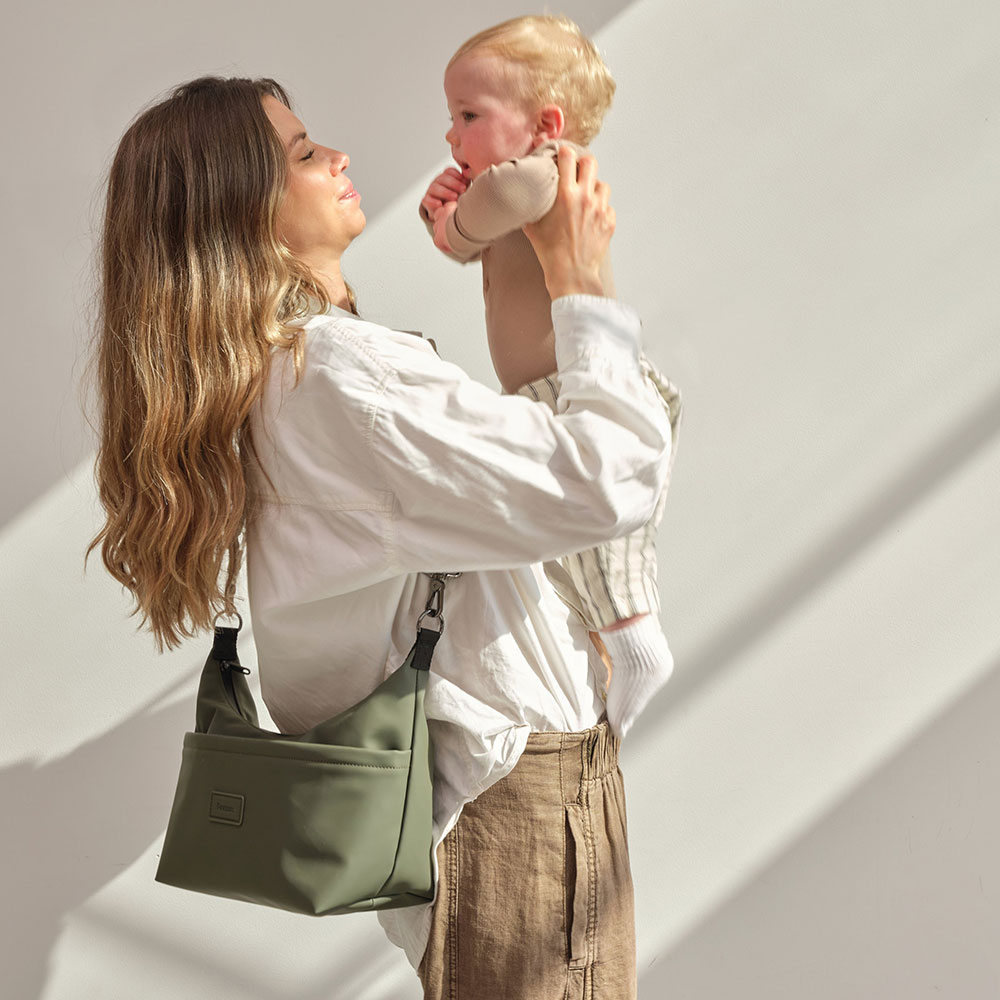 A mother with a khaki green bag holding a baby in the air