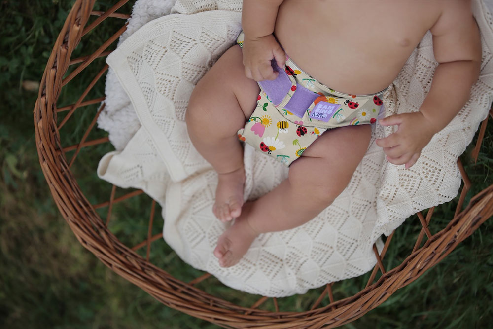A baby laid on a blanket inside a wicker basket wearing a nappy with nature print