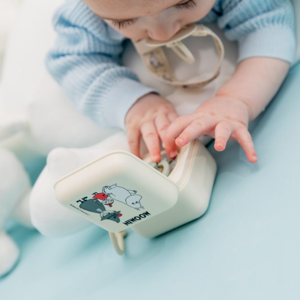 A baby lying on a blue surface with a BIBS pacifier reaching for a pacifier box from the BIBS x Moomin collaboration 