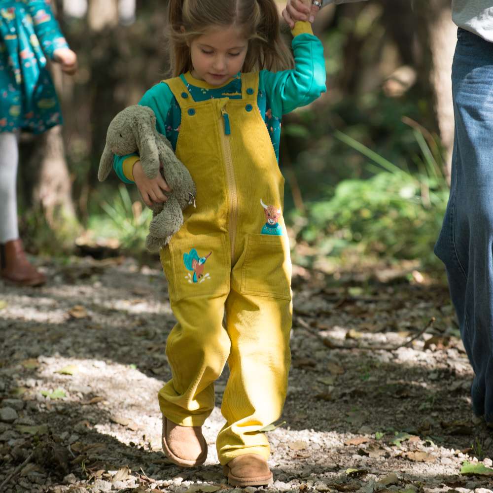 A child walking outside holding an adult's hand wearing yellow dungarees and a turquoise top