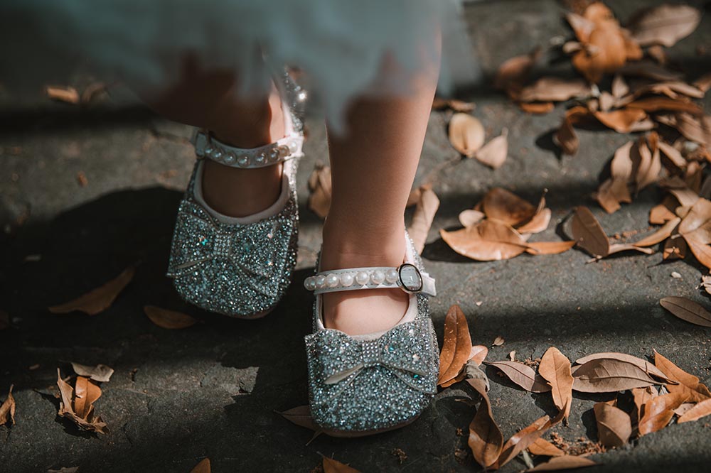 A child's feet shown walking outside among leaves wearing silver sparkly bow shoes with a pearl strap