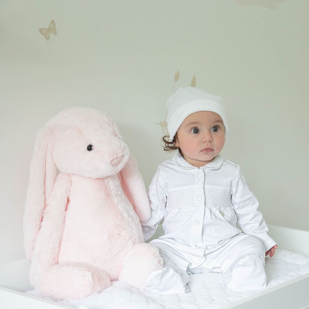 A baby in a white babygro and hat sat beside a large pink rabbit soft toy 