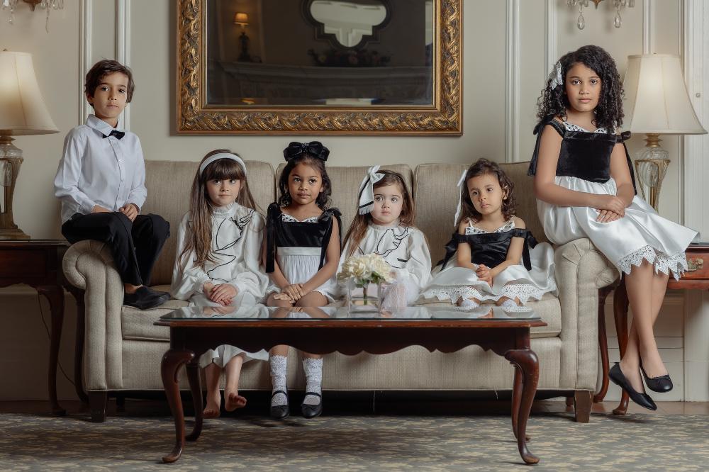 A group of children sat on a sofa in an ornate room wearing black and white occasionwear by Petite Maison Kids