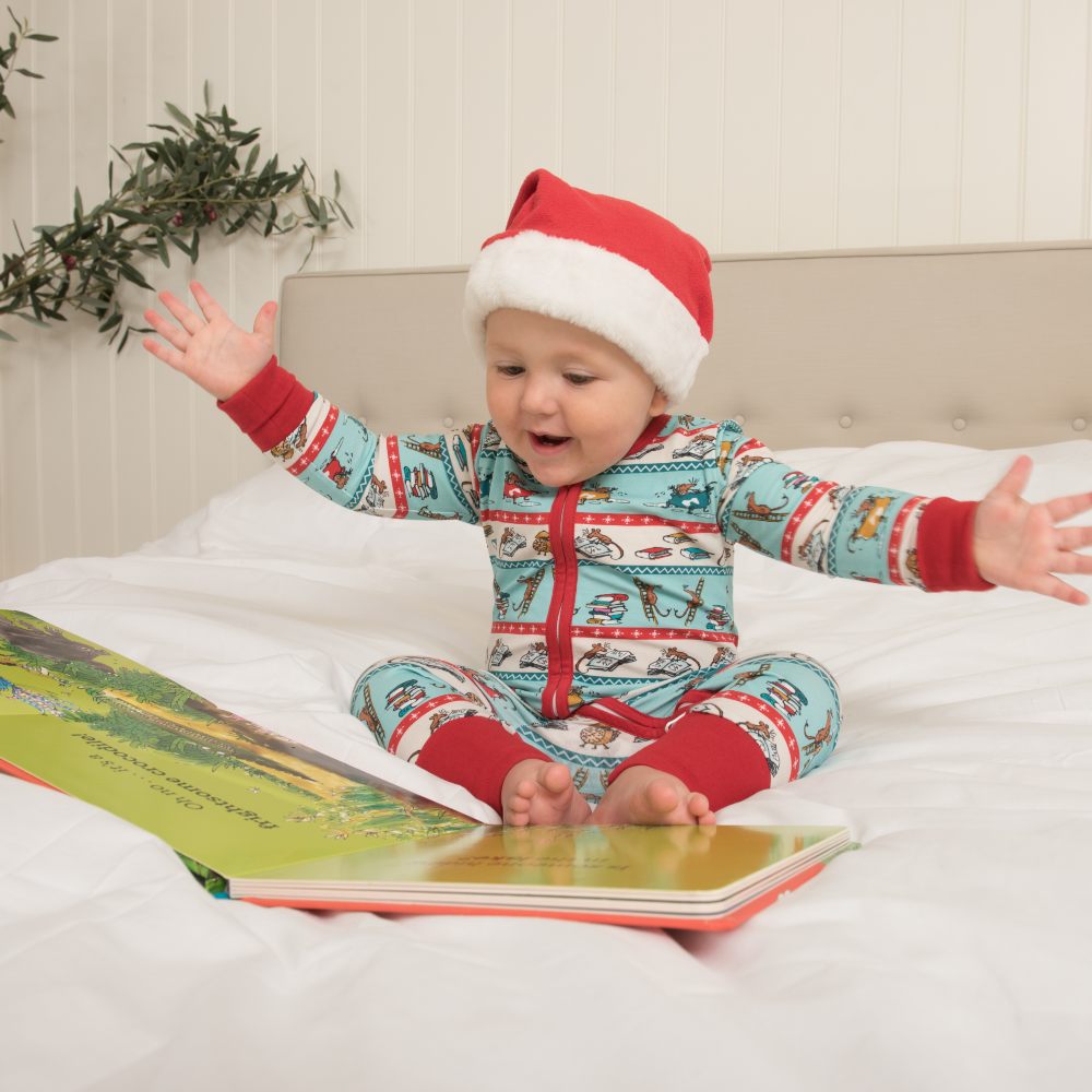 A baby sat on a bed with a large book wearing Roald Dahl print pyjamas by Ducky Zebra and a Christmas hat 