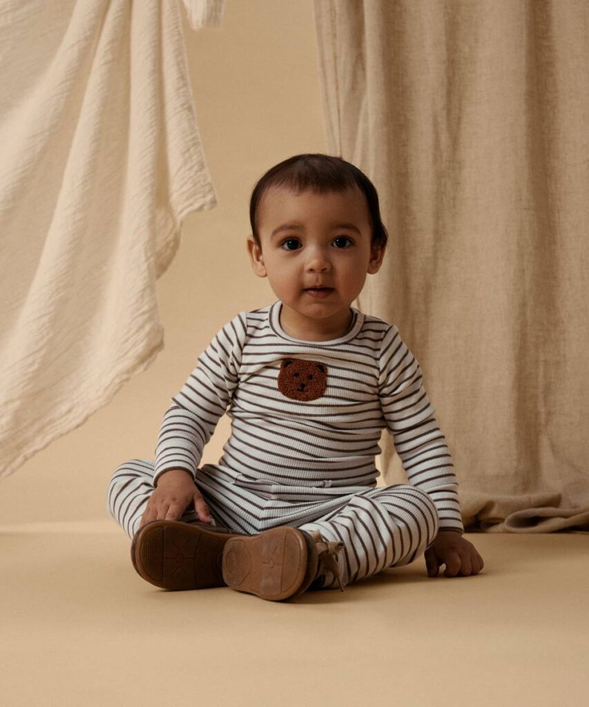 A baby sat on the floor wearing a striped outfit with a teddy bear motif on the front by Bestseller 