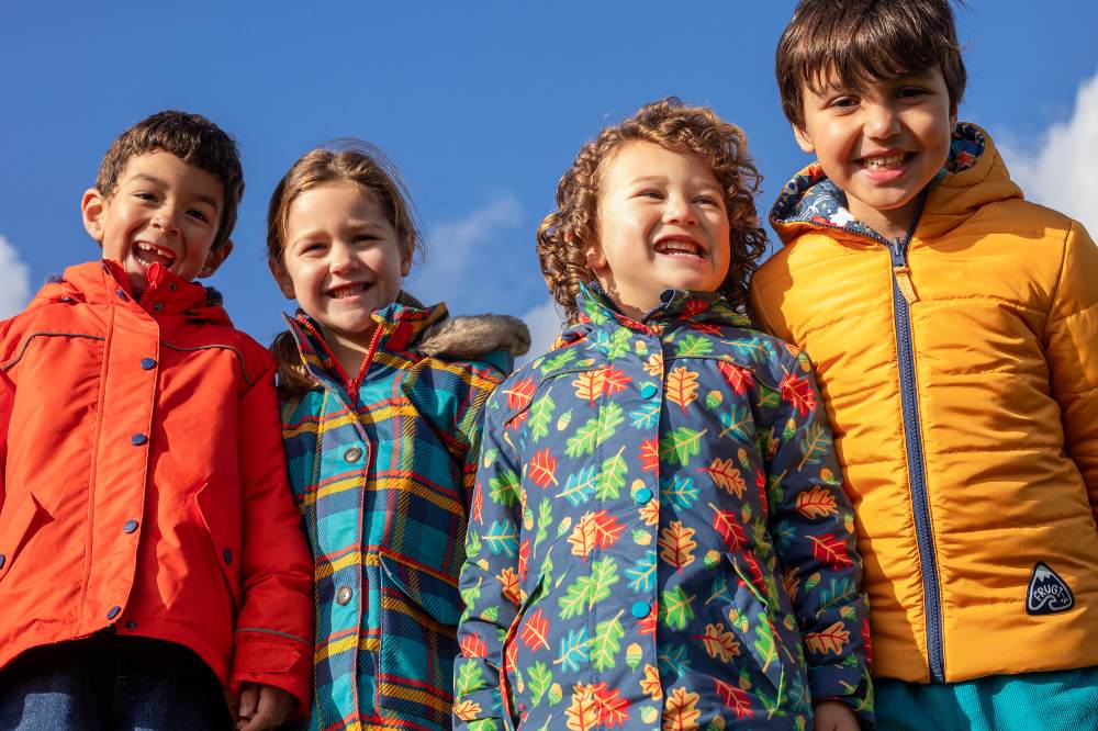 Four children stood together outside against a blue sky wearing winter coats by Frugi