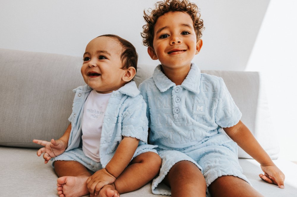 Two young boys smiling on a sofa wearing pale blue outfits by Picture Book Fashion